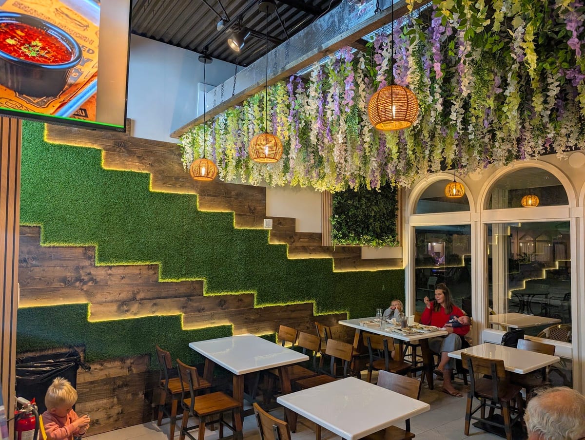 Guests at marble tables beside the green wall and stepped wood, under hanging wisteria and woven lamps